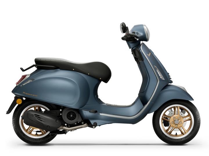 The Vespa Primavera 125 Officina 8 Euro 5+ A blue Vespa scooter with a rounded design and gold accents, viewed from the side, showcasing its seat, handlebars, and wheels.