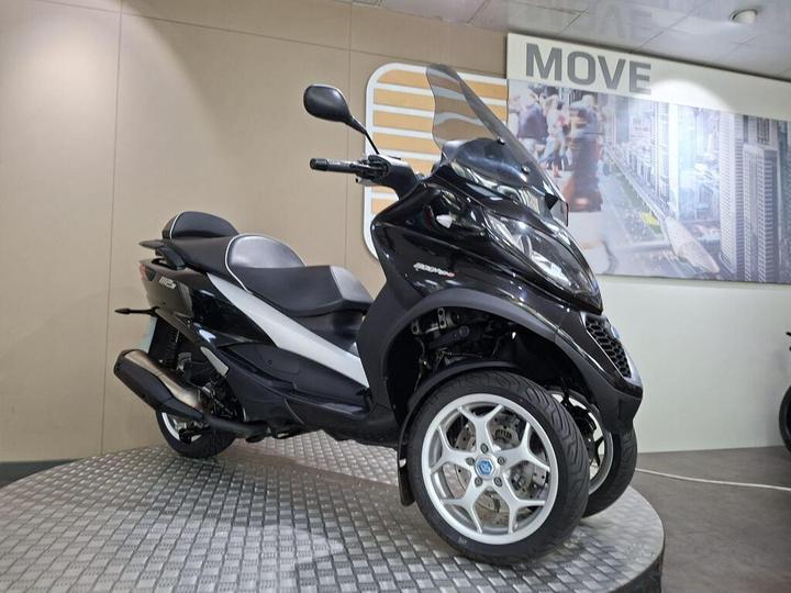 A black and white three-wheeled scooter displayed on a circular platform, with a modern backdrop featuring the word "MOVE" and a cityscape. The scooter has a streamlined design and large alloy wheels.