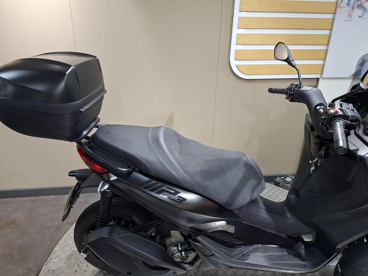 A black scooter with a covered seat and a matching rear storage box, positioned in a modern indoor setting with a neutral background.