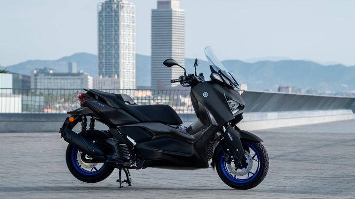 A sleek black Yamaha XMAX scooter is parked on a flat surface with a city skyline in the background, featuring modern buildings and mountains in the distance.