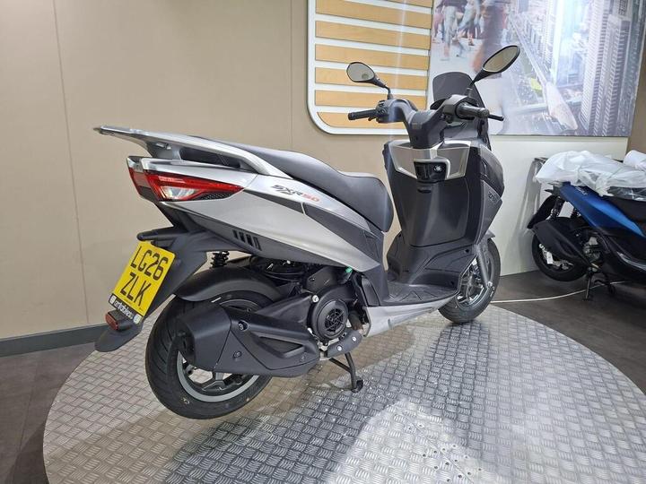A modern silver and black scooter displayed on a circular metallic platform in a showroom, with a blurred background featuring a cityscape and another scooter partially visible. The scooter has a license plate visible.