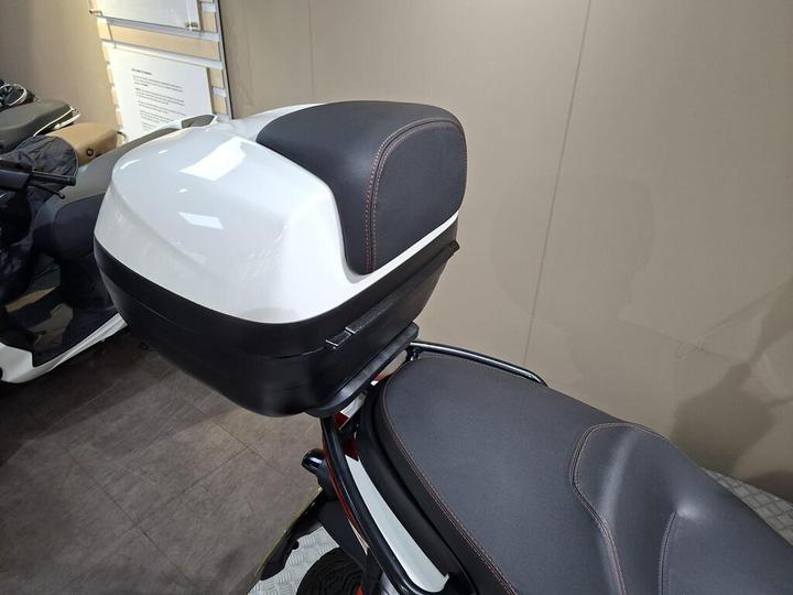 A close-up view of a white scooter top box mounted on the rear seat, featuring a sleek design with black accents and stitching.