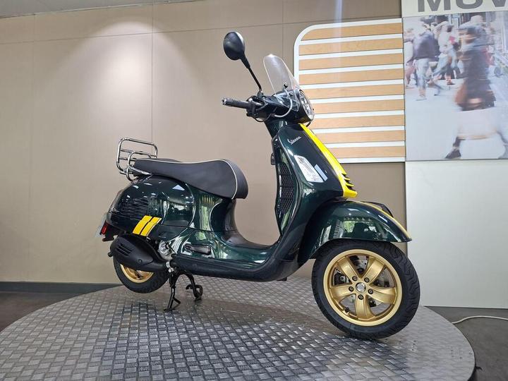 A green and yellow Vespa scooter on a circular display stand, positioned against a neutral background with soft lighting. The scooter features gold alloy wheels and a rear luggage rack.