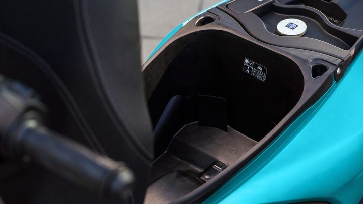 Close-up of the storage compartment of a scooter, featuring a textured black interior and a circular cover with a logo, alongside printed specifications on one side. The exterior is a bright turquoise color.