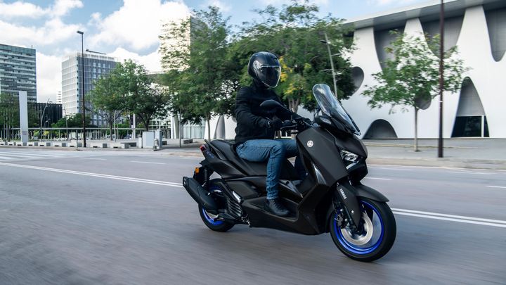A person wearing a black helmet and jacket is riding a sleek black scooter with blue wheels on a city street lined with modern buildings and trees.