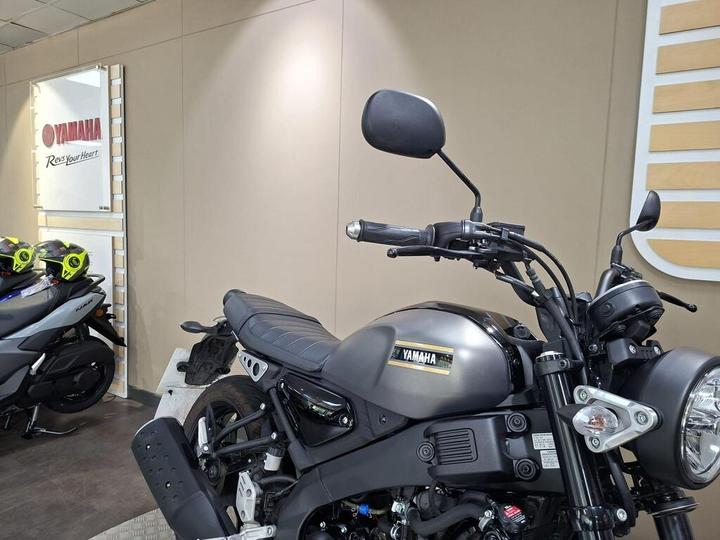 A close-up of a Yamaha motorcycle showcased in a store, with a sleek black body and a visible logo on the tank. Behind the motorcycle, there is a light-colored wall with a Yamaha sign and two helmets on the floor.