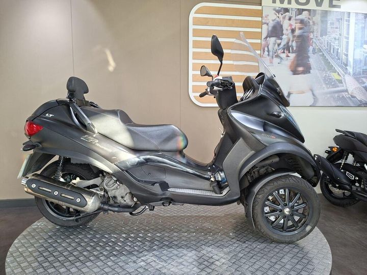Piaggio MP3 500 Sport Touring LT (2017) A black three-wheeled scooter displayed on a textured circular platform, with a modern backdrop featuring a city scene. The scooter has a windshield and a sleek design, showcasing its sporty aesthetics.
