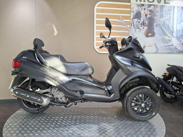 Piaggio MP3 500 Sport Touring LT (2017) A black three-wheeled scooter displayed on a textured circular platform, with a modern backdrop featuring a city scene. The scooter has a windshield and a sleek design, showcasing its sporty aesthetics.