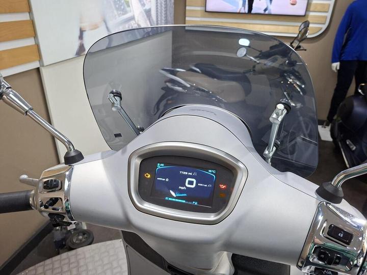 Close-up view of a scooter's dashboard, featuring a digital display showing speed, temperature, and warning indicators, with a clear windshield and handlebars.