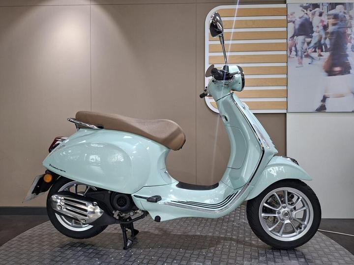 A teal-colored scooter with a brown seat displayed in a showroom, featuring chrome accents and a modern design.
