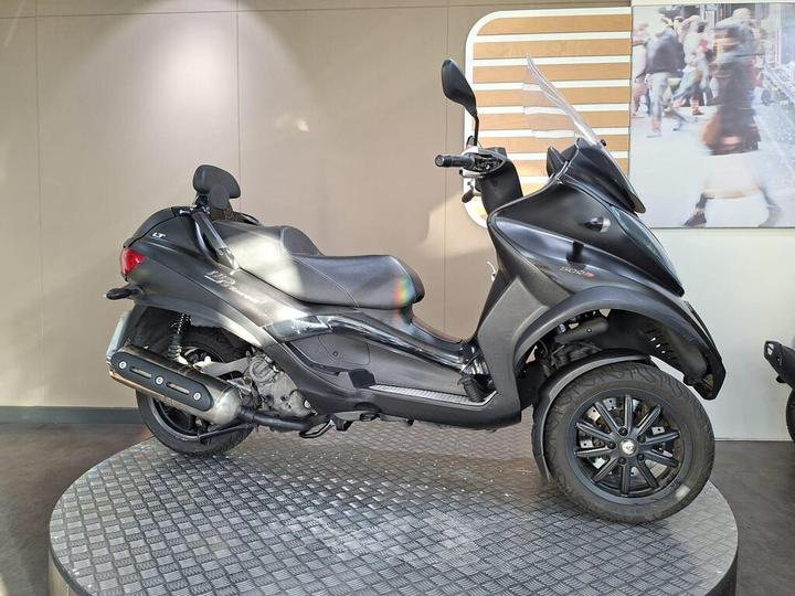 A black scooter displayed on a circular platform against a neutral background, showcasing its sleek design and detailing.