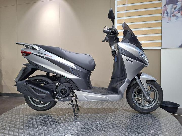 Aprilia SXR 50 2026 A silver Aprilia scooter displayed on a textured surface against a neutral background.