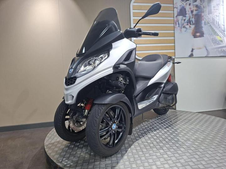 A compact three-wheeled scooter with a sleek silver and black design, featuring a large windshield and dual front wheels, displayed on a circular metallic platform against a neutral background.