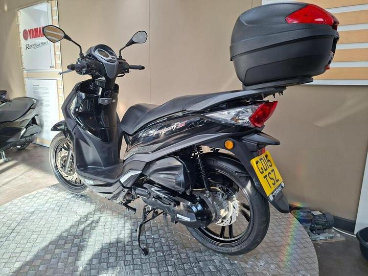 A black scooter with a top box is displayed on a patterned gray surface, showcasing its side profile and sleek design, against a light-colored wall featuring Yamaha branding.