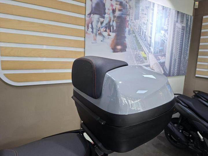A gray motorcycle top case with a black cushioned backrest is displayed against a backdrop of a cityscape with people walking, inside a showroom.