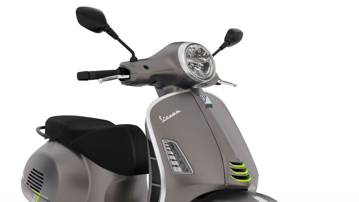 A close-up view of a gray Vespa scooter, featuring a round headlight, black handlebars, and a sleek design with green accents on the body and seat.