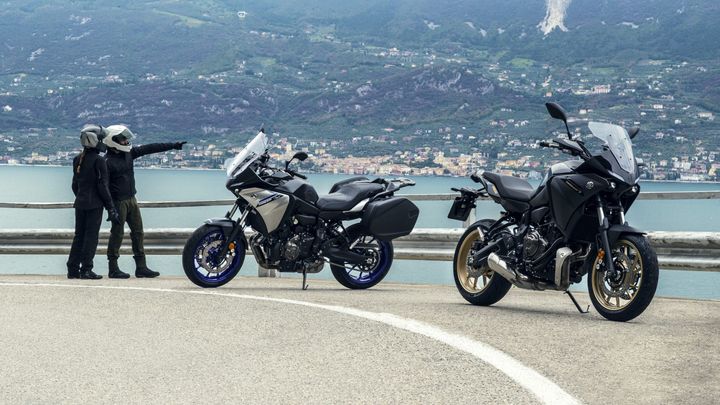 Two motorcyclists, one pointing towards the landscape, stand beside two sport-touring motorcycles parked on a winding road overlooking a lake and mountains.