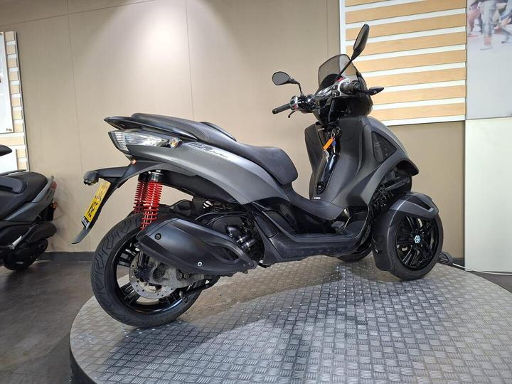 A black and gray scooter with a sleek design, featuring a rear shock absorber in red, positioned on a circular platform with a textured surface, against a neutral wall backdrop.