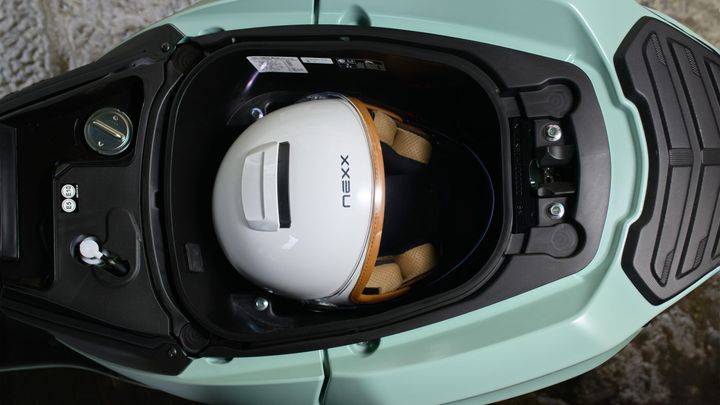 An overhead view of a light green scooter storage compartment featuring a white Nexx motorcycle helmet and various small components, including a tank cap and a socket.