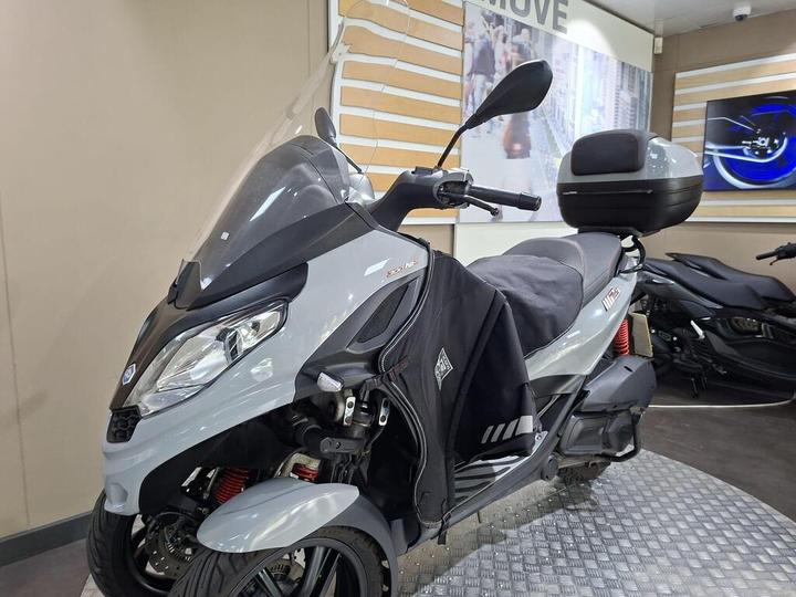 A grey scooter with a windshield, large rear storage box, and a black seat cover, displayed in a modern showroom with a textured floor.
