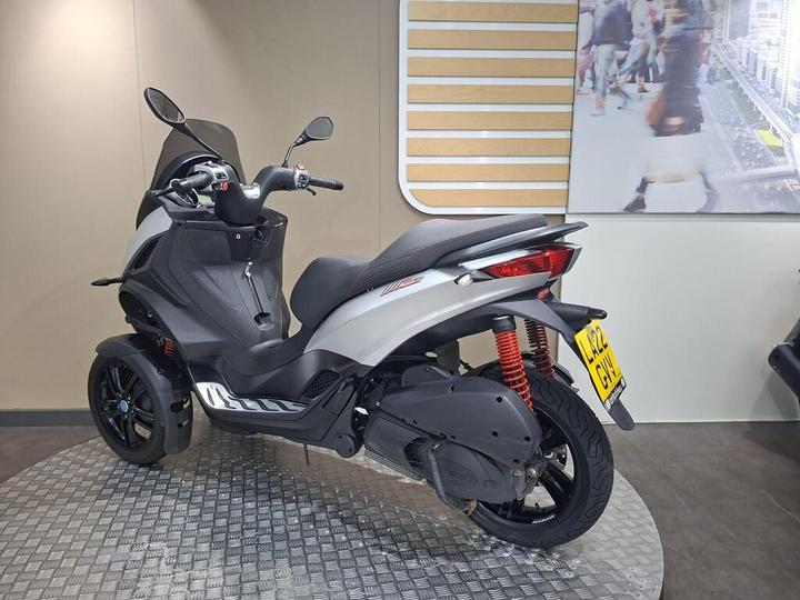 A sleek, modern three-wheeled scooter in silver and black with a sporty design, displayed against a neutral background. The rear showcases red shock absorbers and a distinctive wheel design, highlighting its contemporary features.