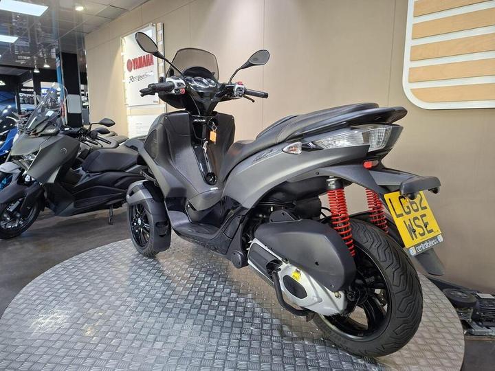 A parked gray scooter with distinctive red suspension springs is displayed on a circular metal platform in a dealership. The background features other scooters and a Yamaha branding sign.