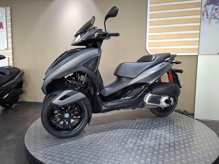 A sleek, modern scooter with a matte gray finish is displayed on a circular platform in a well-lit showroom. The scooter features distinct design elements, including a prominent front shield and stylish black wheels, with red accents on the suspension. In the background, blurred silhouettes of other scooters can be seen against a neutral-colored wall.