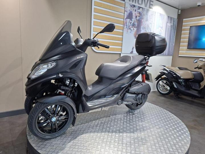 A black three-wheeled scooter displayed in a showroom, featuring a windshield, a comfortable seat, and a top case for storage, with a backdrop of a city-themed wall and another scooter in beige visible in the background.