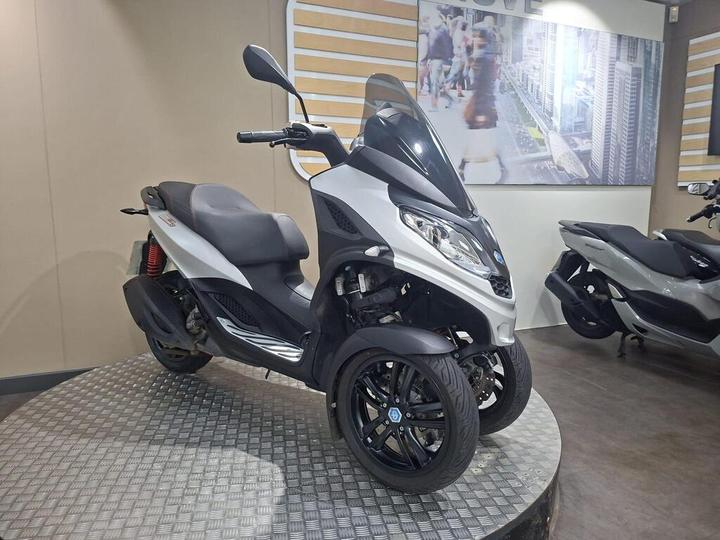 A three-wheeled scooter in a showroom, featuring a sleek silver design with a windscreen and black accents, parked on a circular platform with a textured surface.