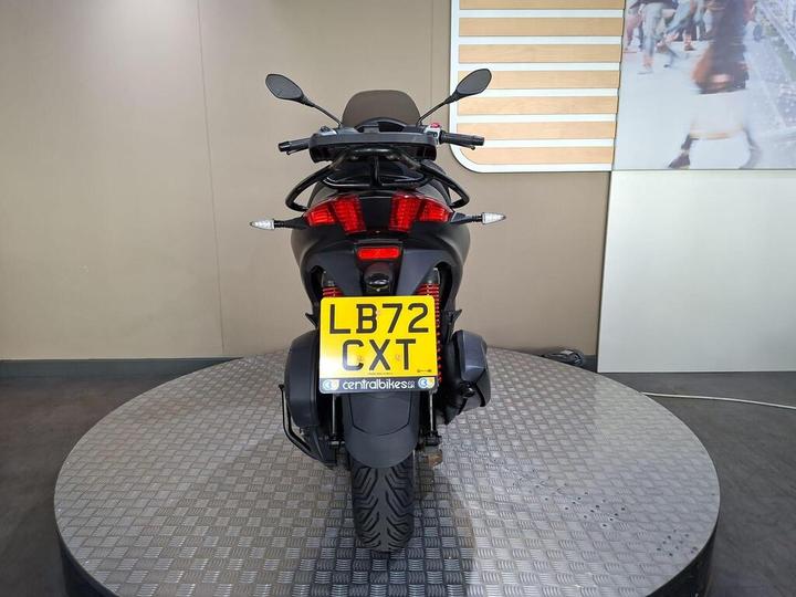 A black motorcycle viewed from the rear, featuring a license plate reading "LB72 CXT" and positioned on a circular display platform with a patterned surface.
