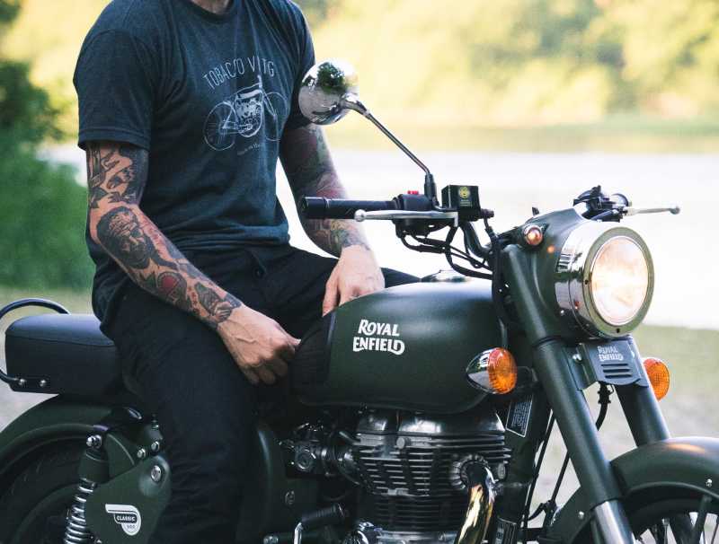 A male with a green shirt and tattoos sitting on a classic green Royal Enfield motorcycle