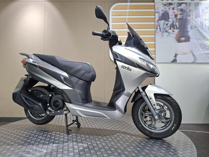 A silver Aprilia scooter displayed on a circular metal platform, with a minimalist background featuring a blurred image of people.