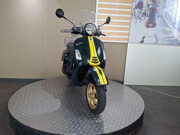 A green Vespa scooter with yellow accents is displayed on a circular metal platform in a showroom setting, featuring a clear windshield and a decorative backdrop.