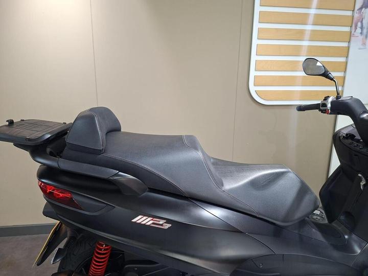 A black scooter seat is shown from the side, featuring a smooth, contoured design with a passenger backrest and a luggage rack. The background is a neutral beige wall, enhancing the focused view of the scooter.