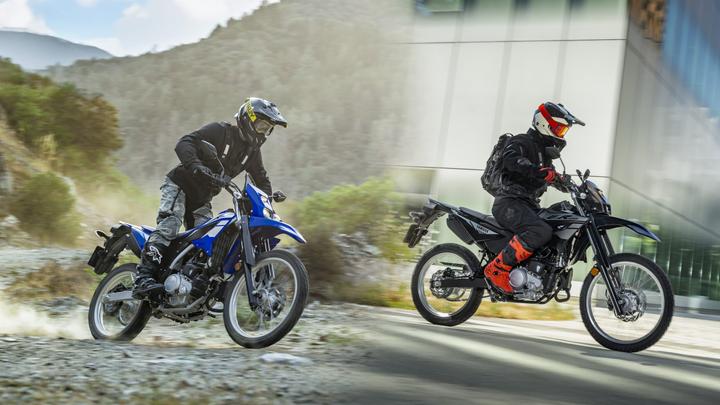A split image shows two riders on dirt motorcycles. On the left, a rider in a black jacket and camouflage pants rides a blue bike on a gravel path, surrounded by greenery and mountains. On the right, a rider in a black suit and red accents rides a black motorcycle along a modern building, with a smooth surface and urban backdrop.