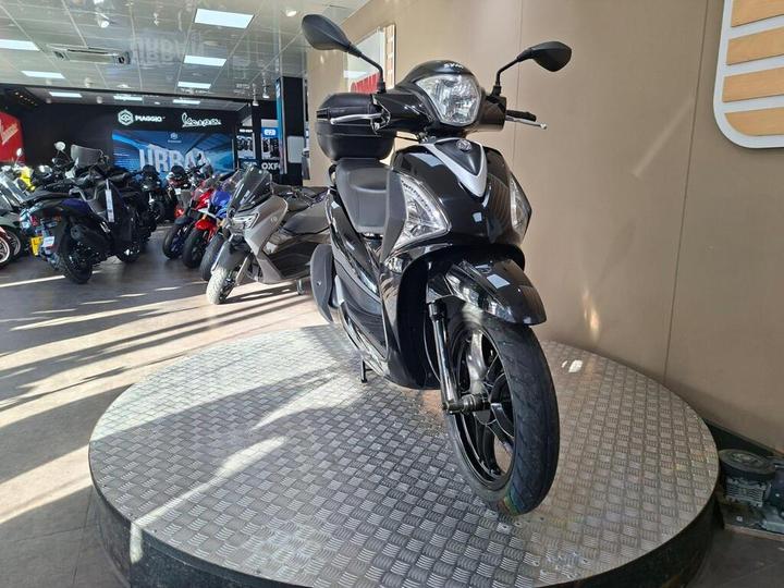 A black scooter displayed on a circular platform in a motorcycle showroom, featuring various bikes in the background and bright overhead lighting.