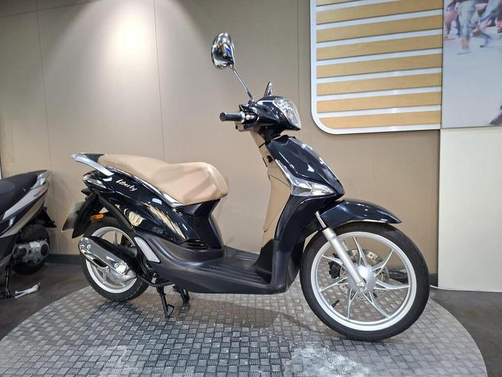 A stylish black scooter with a beige seat on a textured circular platform, set against a neutral wall background with subtle design elements.