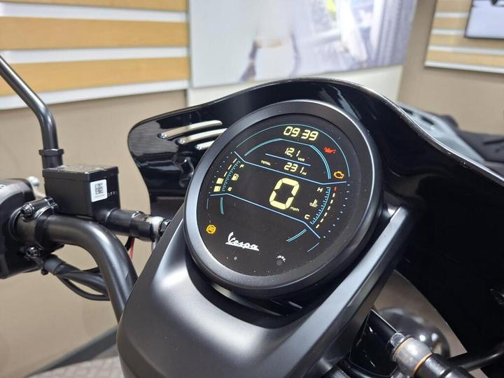 A close-up view of a motorcycle dashboard display showing time (09:39), voltage (12.1V), fuel level (23%), and speed (0 mph), with colorful indicators and a sleek black design.