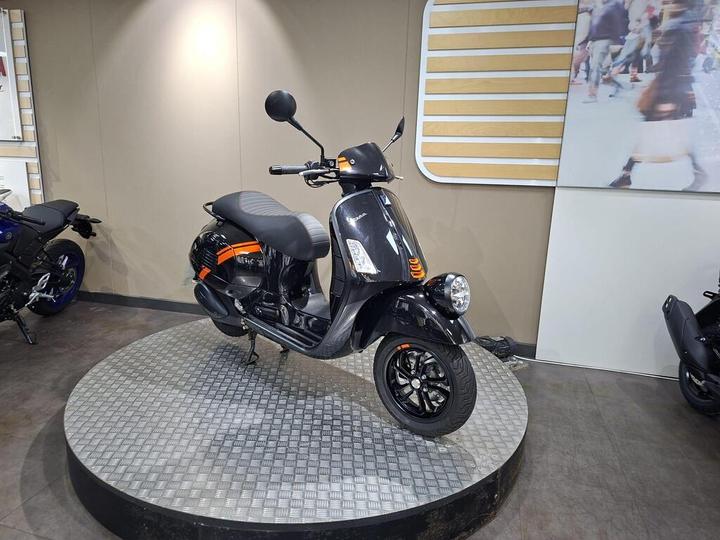 A black scooter with orange accents is displayed on a circular platform in a showroom. The scooter features a sleek design with a low seat and a round headlight. In the background, partially visible, is another motorcycle. The walls of the showroom are neutral in color, enhancing the focus on the scooter.