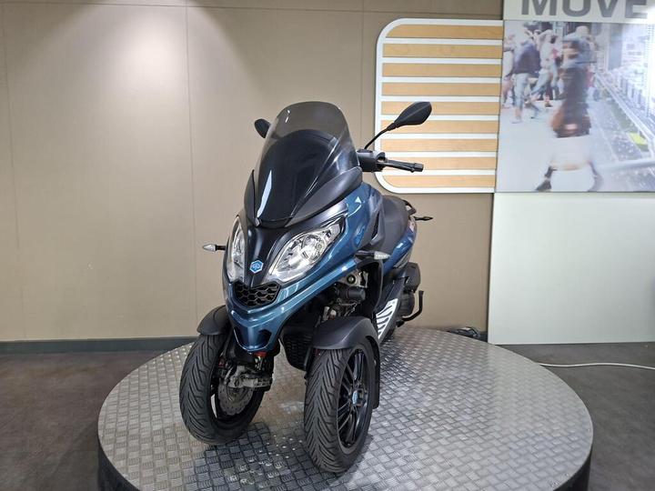 A blue three-wheeled scooter displayed on a rotating pedestal in a showroom, with a neutral background featuring a graphic on the wall.
