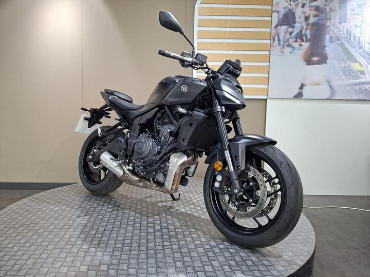 A black motorcycle is displayed on a round platform, with the front handlebars turned slightly to the right. The background features a beige wall and a blurred image of pedestrians.