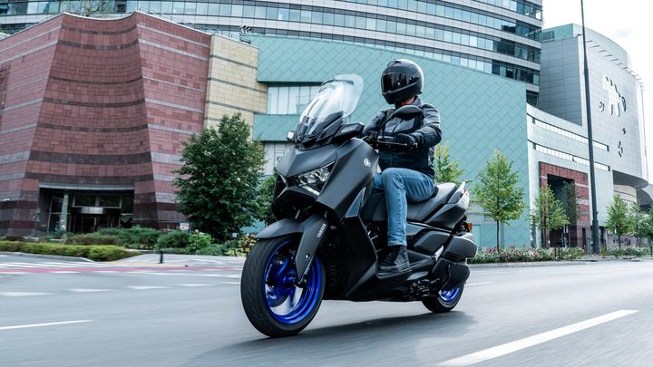 A motorcyclist in a black helmet and jacket rides a matte black scooter with blue wheels, cruising along a city street with modern buildings and greenery in the background.