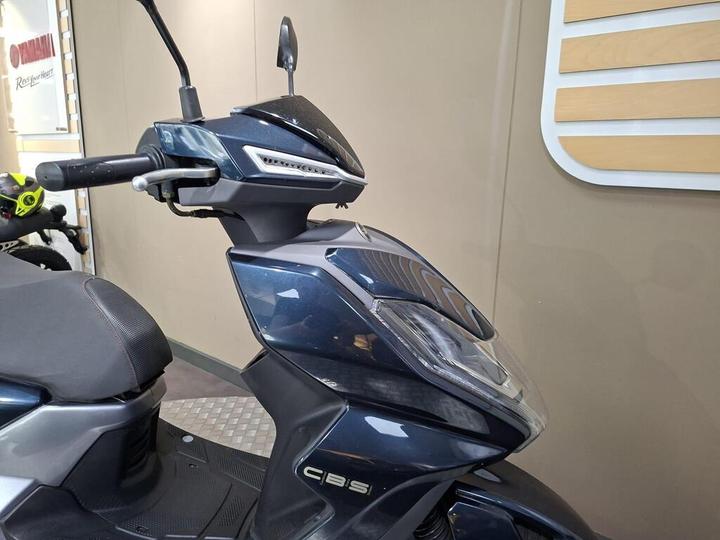 A close-up view of a dark blue scooter's front, featuring the handlebar, headlight, and sleek body design, against a neutral background.