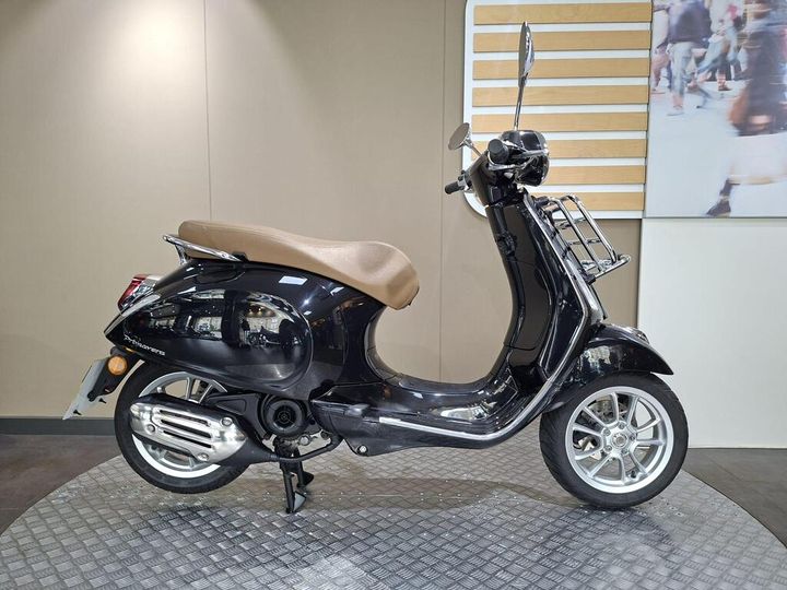 Vespa Primavera 50 (2020) Black scooter with a brown seat displayed on a circular patterned platform against a neutral background.