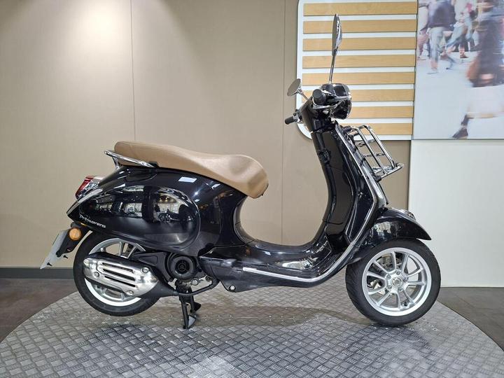 Vespa Primavera 50 (2020) Black scooter with a brown seat displayed on a circular patterned platform against a neutral background.