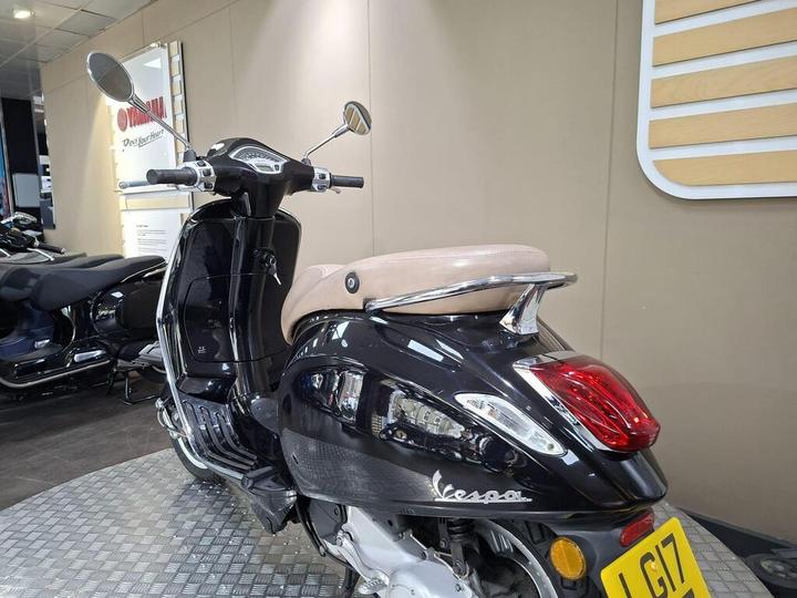 A black Vespa scooter with a tan seat is prominently displayed in a showroom with soft lighting and a backdrop of motorcycles. The scooter features chrome mirrors and a distinctive logo on the side.