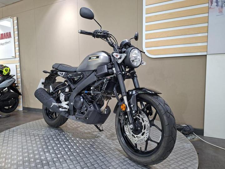 A Yamaha motorcycle displayed on a textured circular platform in a showroom, featuring a sleek black design, a round headlight, and a prominent exhaust.