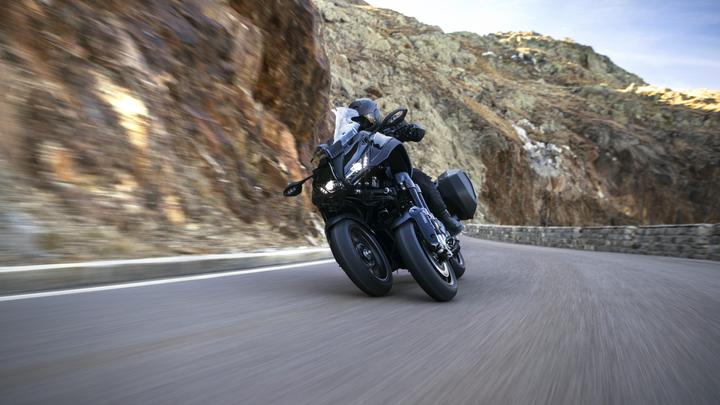 A motorcycle leans into a turn on a winding road surrounded by rocky hills, with motion blur indicating speed.