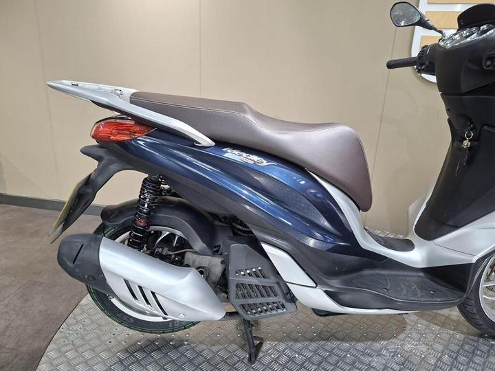 A side view of a blue and silver scooter featuring a brown seat, standing on a checkered floor with a neutral background.