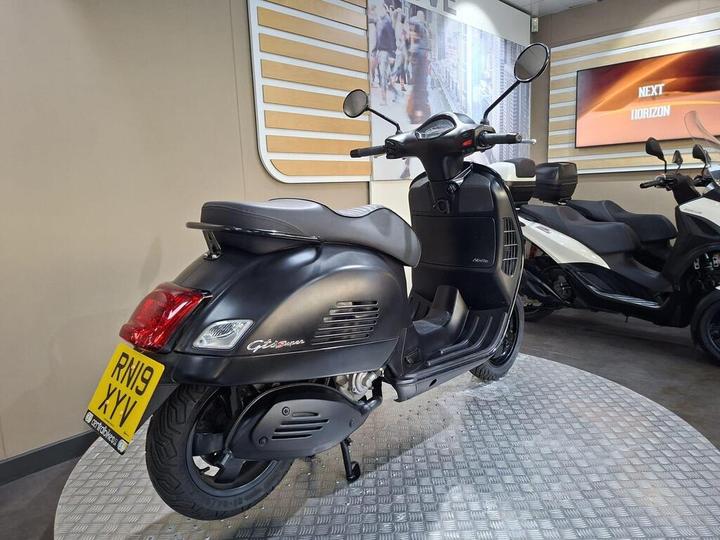 A black Vespa GTS Super scooter is displayed in a showroom setting, showcasing its sleek design, with a license plate reading "RN19 XYV" visible. The background features a modern interior with promotional screens and other scooters.